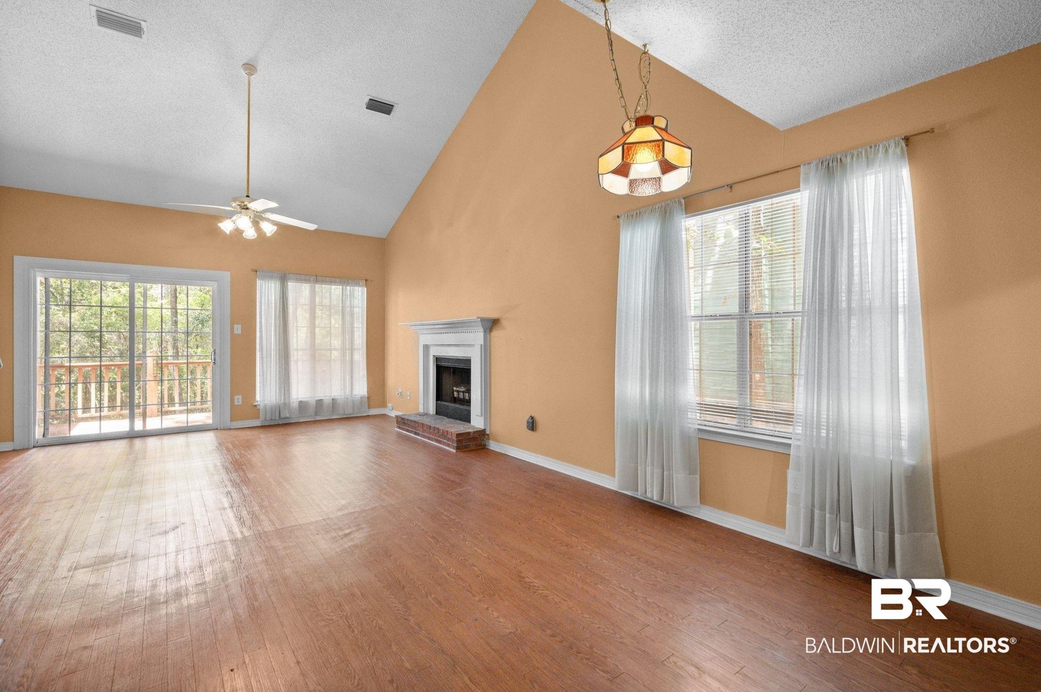 Image 4: Spacious living area featuring a vaulted ceiling with a fan, wood-finish flooring, a fireplace with a white mantel and brick hearth, and multiple windows providing natural illumination