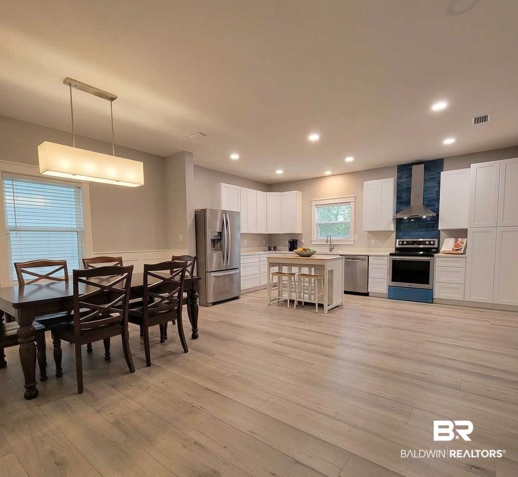 Image 4: Open-concept kitchen and dining area featuring light wood-finish flooring, white cabinetry, stainless steel appliances, and a blue glass tile backsplash to bring a feeling of coastal living to your recipes