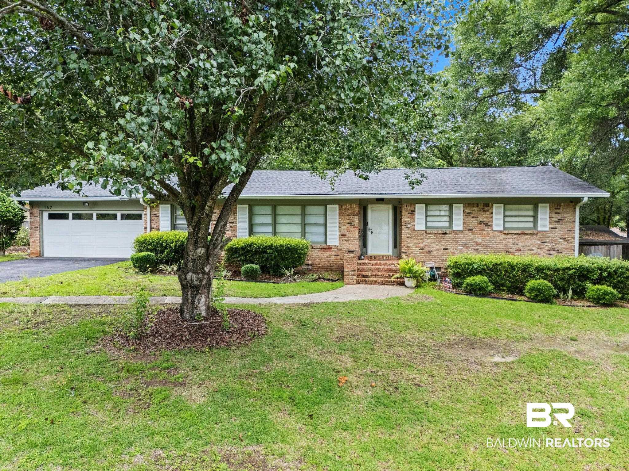 Image 1: Brick-sided residence featuring a prominent mature tree in the front yard, a white paneled garage door, a front entry with brick steps, a dark shingle roof, and white-framed windows with decorative shutters