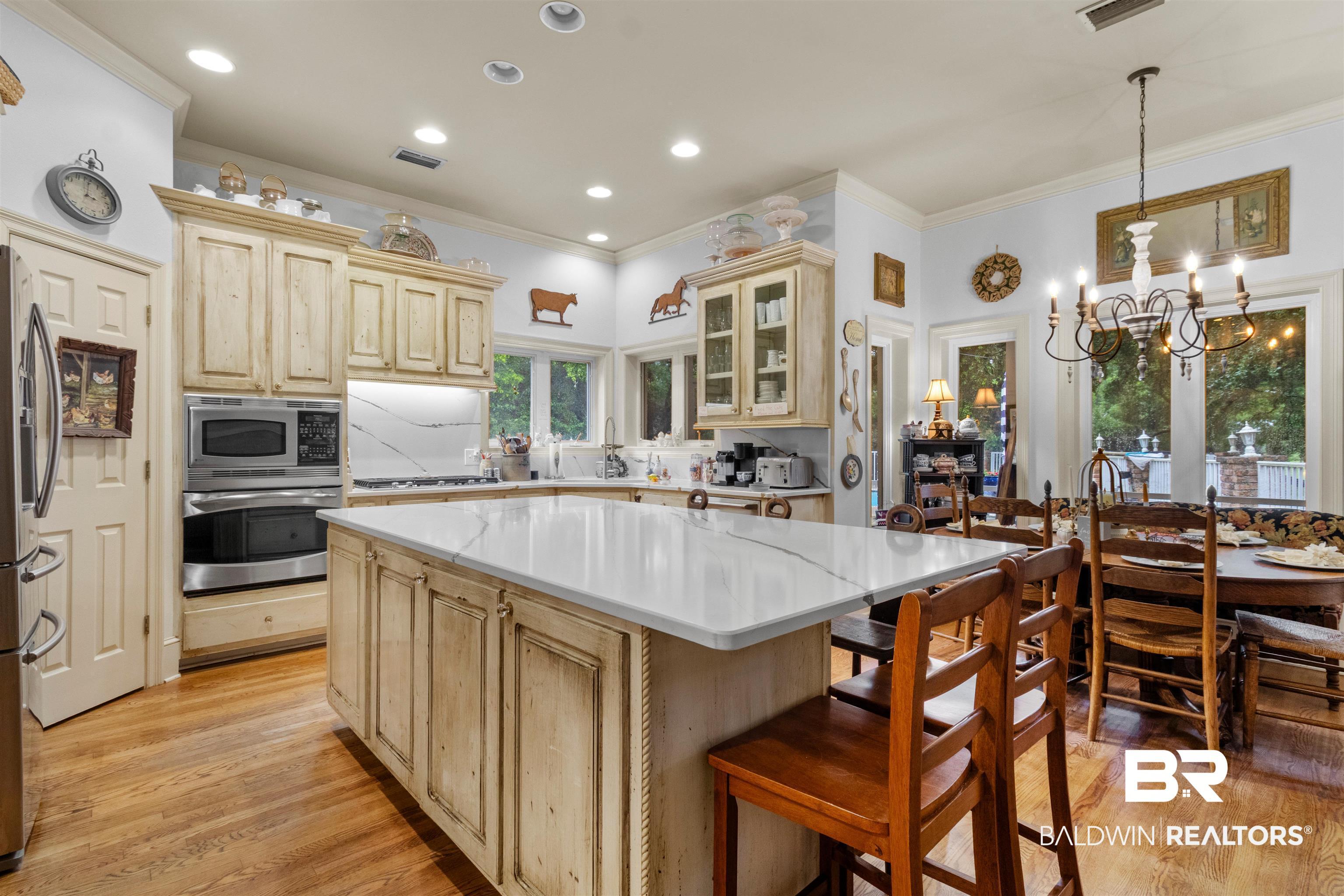Image 3: Kitchen featuring a substantial central island with a light-toned countertop, wood-finish flooring, recessed lighting, built-in stainless steel appliances, and custom cabinetry