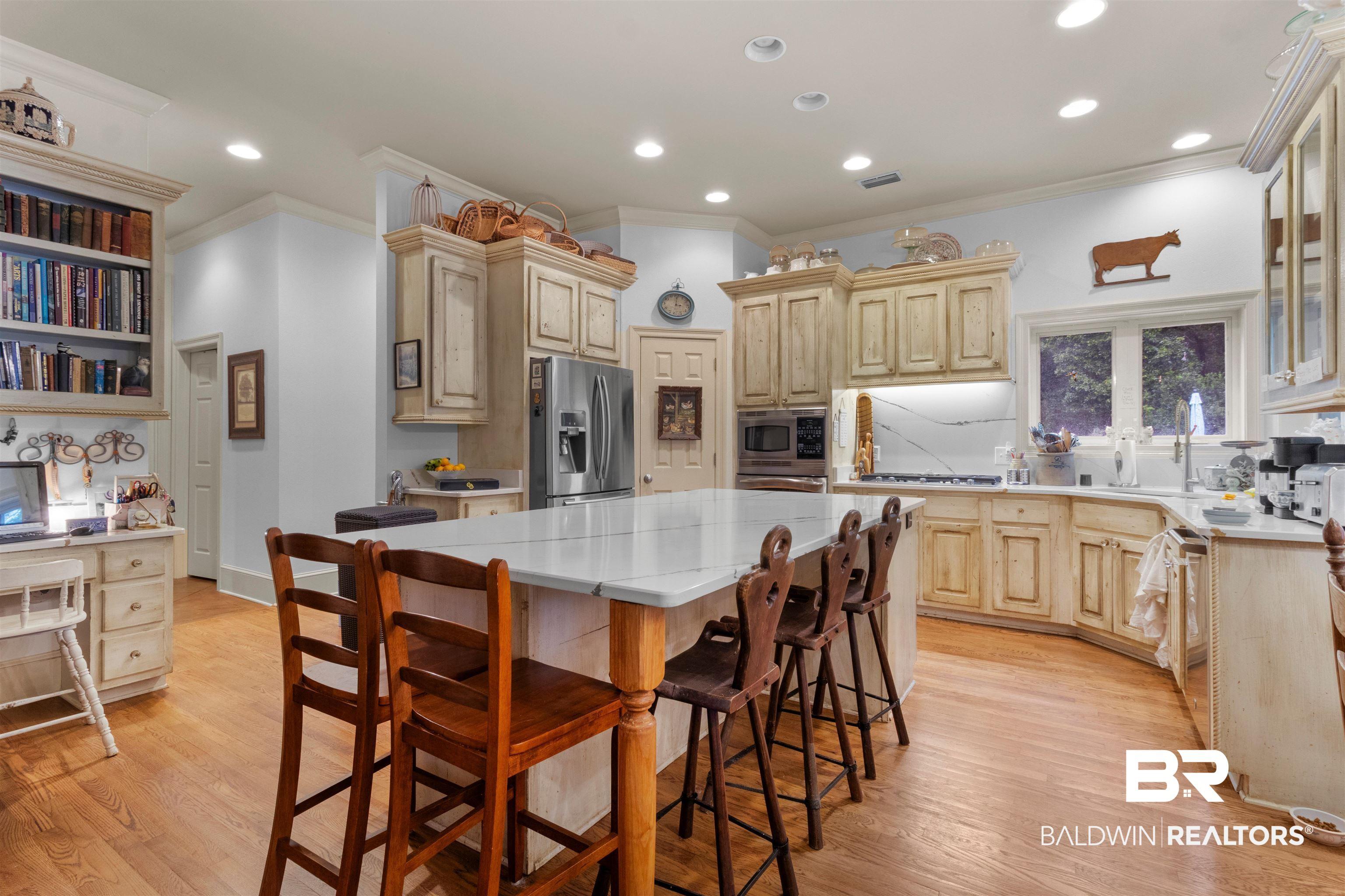 Image 4: Expansive kitchen featuring a large central island with seating, wood-finish flooring, custom cabinetry, stainless steel appliances, and built-in desk with shelving