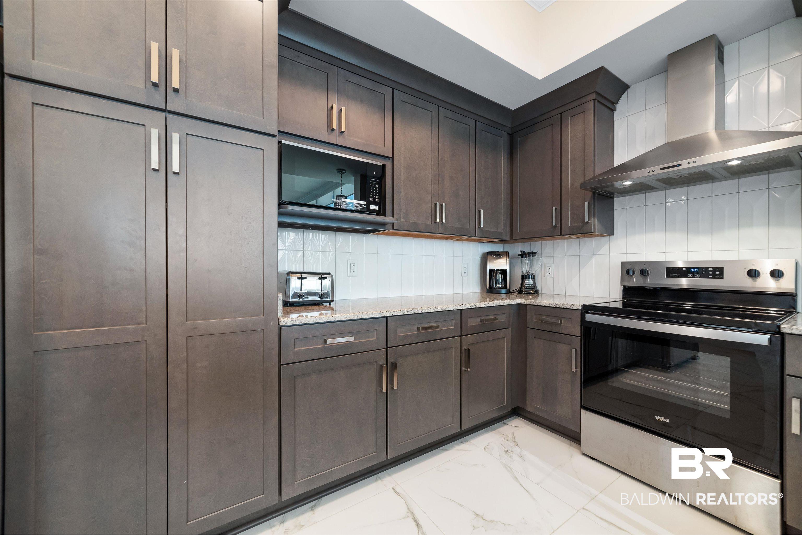 Image 4: Modern kitchen featuring dark wood-finish cabinetry, stainless steel appliances, granite countertops, a subway tile backsplash, and large format tile flooring