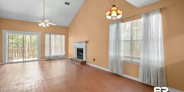 Image 4: Spacious living area featuring a vaulted ceiling with a fan, wood-finish flooring, a fireplace with a white mantel and brick hearth, and multiple windows providing natural illumination