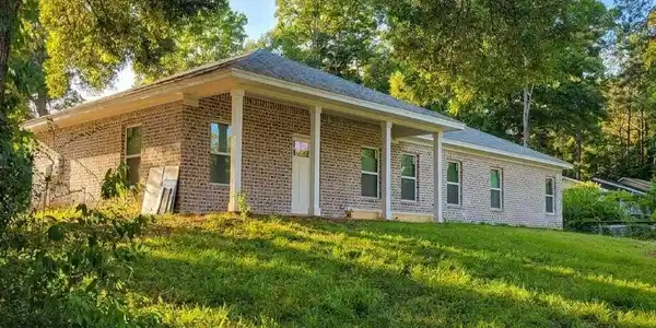 Image 2: Brick exterior home featuring a covered front porch with white columns, multiple windows, and a surrounding natural landscape with mature trees