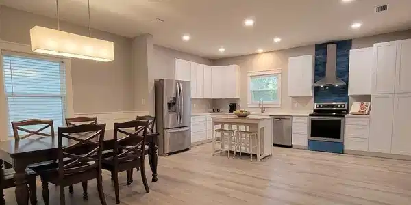 Image 4: Open-concept kitchen and dining area featuring light wood-finish flooring, white cabinetry, stainless steel appliances, and a blue glass tile backsplash to bring a feeling of coastal living to your recipes
