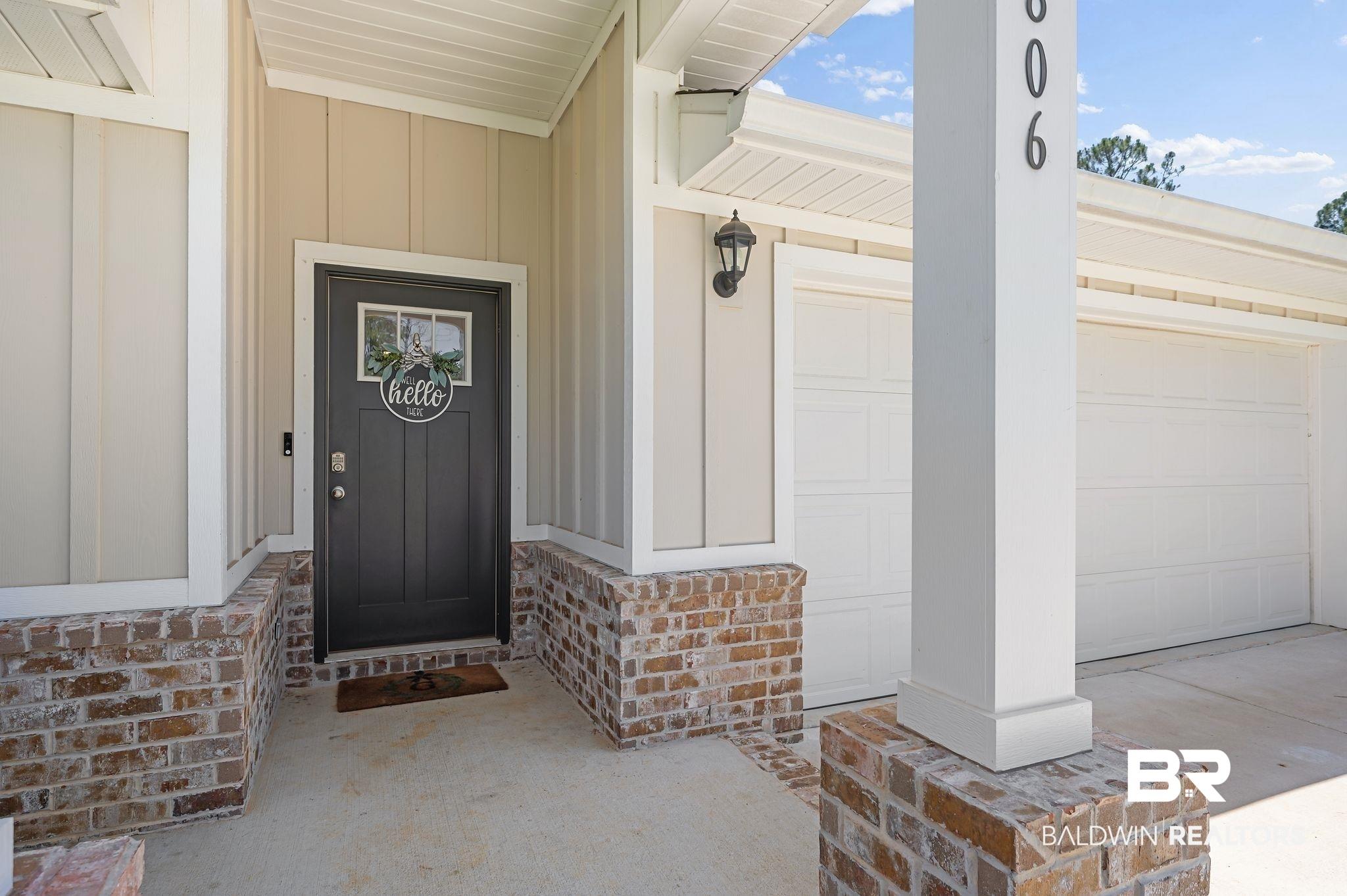 Image 2: Inviting front entry featuring a dark gray door with transom window, board and batten siding, brick skirting, a covered porch, and an exterior wall lantern