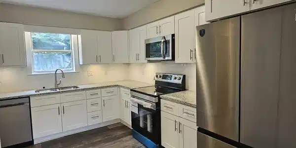 Image 3: Modern kitchen featuring white shaker cabinetry, stainless steel appliances, speckled countertops, wood-finish flooring, and under-cabinet lighting