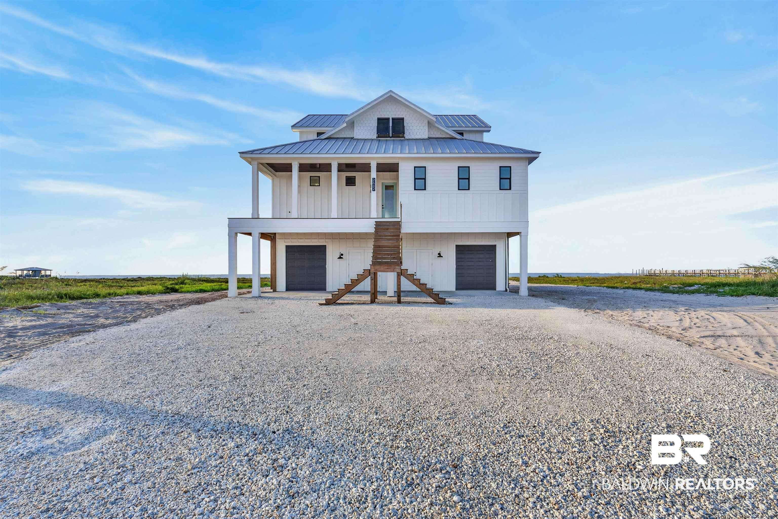 Image 3: Raised coastal-style home featuring white board and batten siding, a metal roof, an elevated covered porch, dual garage bays, and a prominent exterior staircase