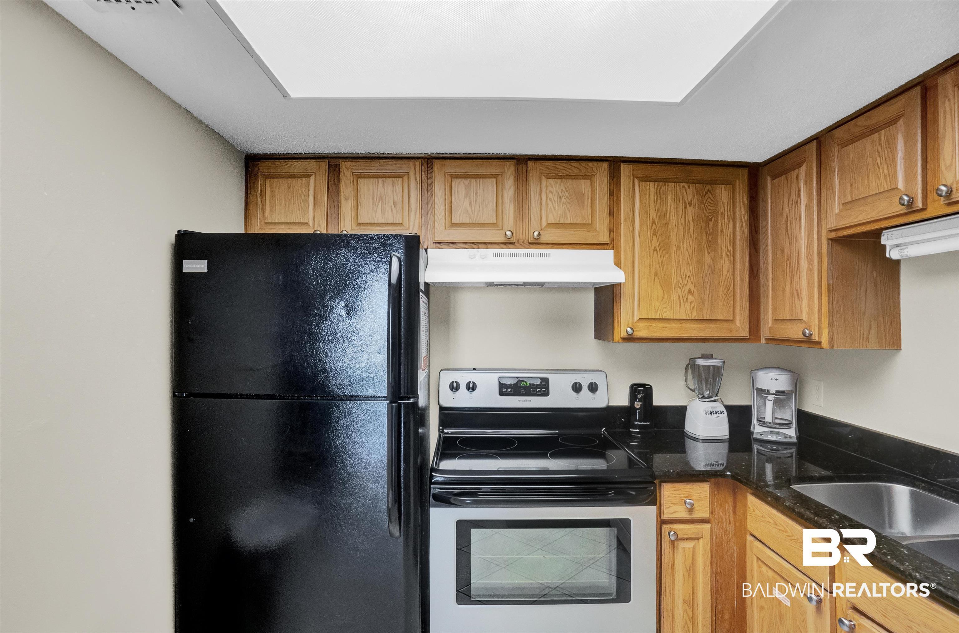 Image 3: Kitchen featuring under cabinet range hood, dark stone countertops, stainless steel range with electric cooktop, brown cabinets, and freestanding refrigerator