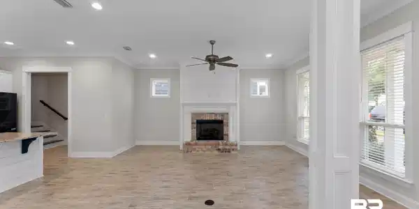 Image 4: Spacious living area featuring light-toned wood-finish flooring, a brick fireplace with a shiplap surround, multiple windows with blinds, a ceiling fan, and recessed lighting