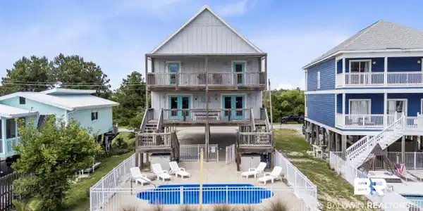 Image 3: Elevated residence featuring a fenced in-ground pool, two levels of covered balconies with teal doors, a gable roof, and wood decking