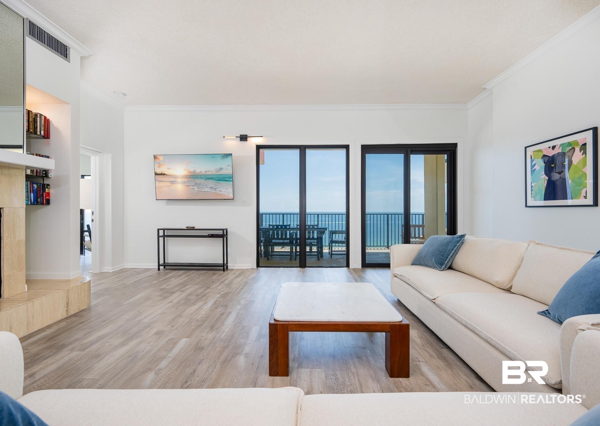 Image 2: Spacious living area featuring light wood-finish flooring, a built-in shelving unit, a wall-mounted flat-screen television, and expansive sliding glass doors opening to a balcony with water scenery