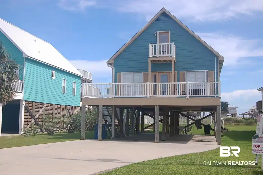 Image 1: Elevated home featuring teal horizontal hardy board siding and a large front balcony with composite decking and white vinyl railing. Front balcony is on the primary level and has an upper-level Juliet balcony from the upstairs bedroom.