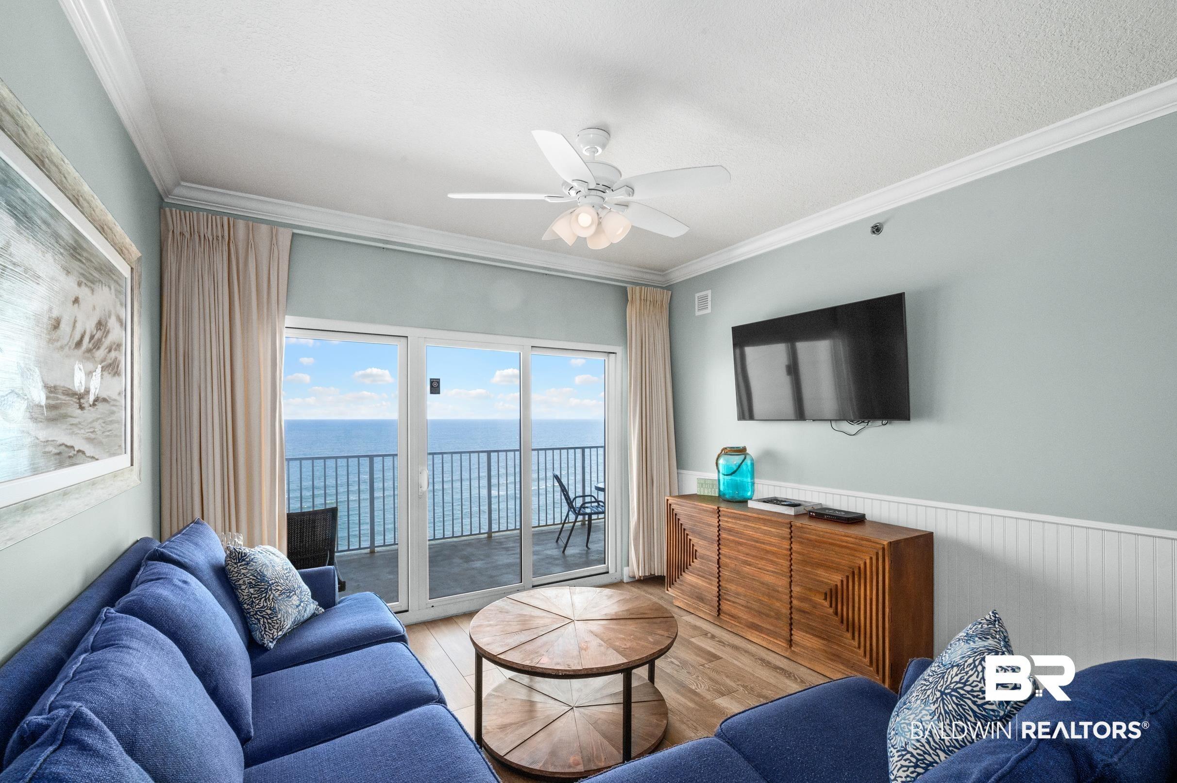 Image 4: Living area featuring a spacious balcony with ocean scenery, wood-finish flooring, white crown molding, and a ceiling fan with integrated lighting