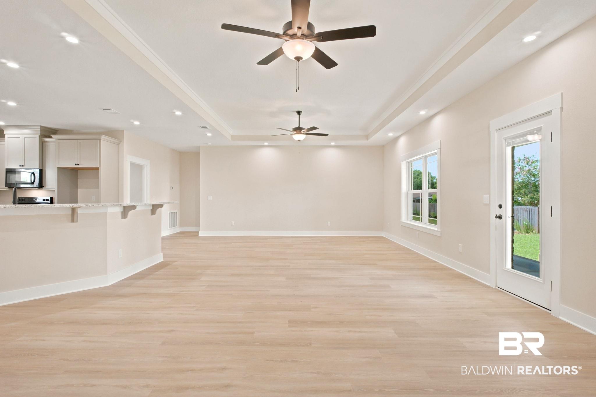 Image 4: Spacious living area featuring light wood-finish flooring, a tray ceiling with recessed lighting and ceiling fans, and a kitchen pass-through with a light-colored countertop