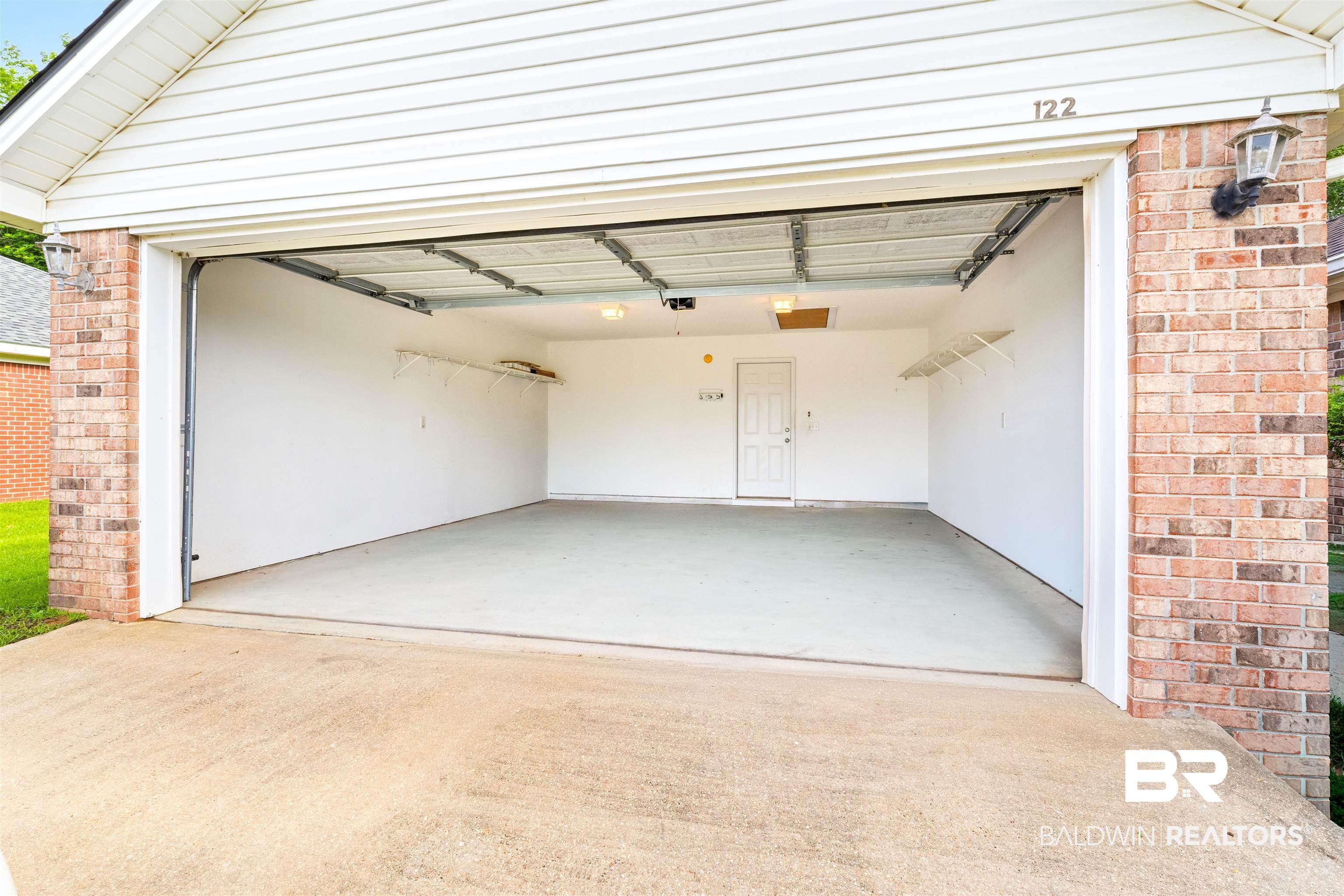 Image 4: Two-car garage with a concrete floor, interior access door, wall-mounted shelving, and overhead lighting