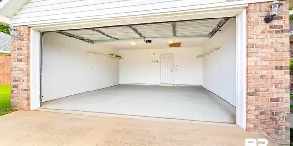 Image 4: Two-car garage with a concrete floor, interior access door, wall-mounted shelving, and overhead lighting