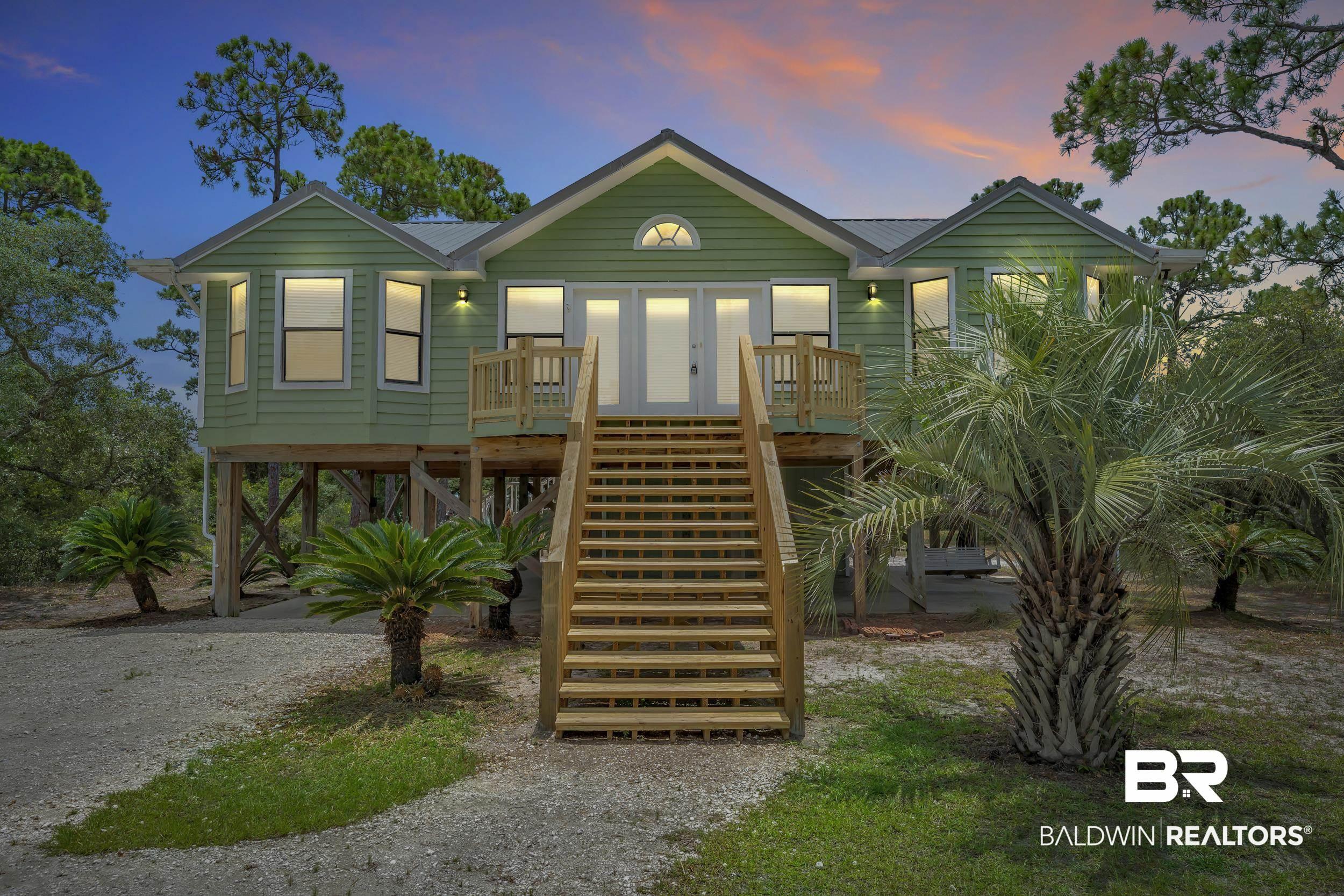 Image 3: Stilt home featuring light green siding, a metal roof, prominent front stairs, a small balcony, and surrounding mature trees