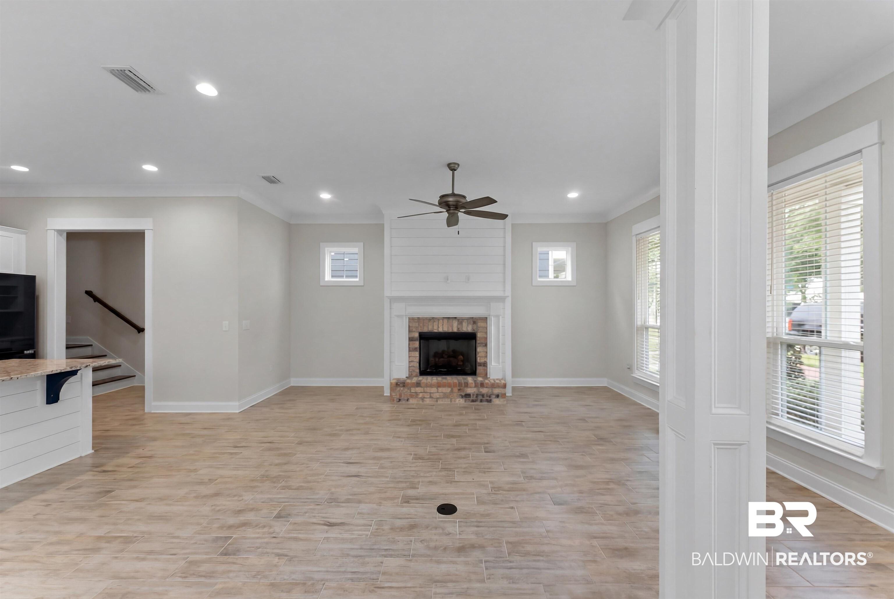 Image 4: Spacious living area featuring light-toned wood-finish flooring, a brick fireplace with a shiplap surround, multiple windows with blinds, a ceiling fan, and recessed lighting