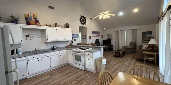 Image 3: Open-concept interior featuring wood-finish flooring, vaulted ceilings with recessed lighting, and a kitchen with white cabinetry and light countertops