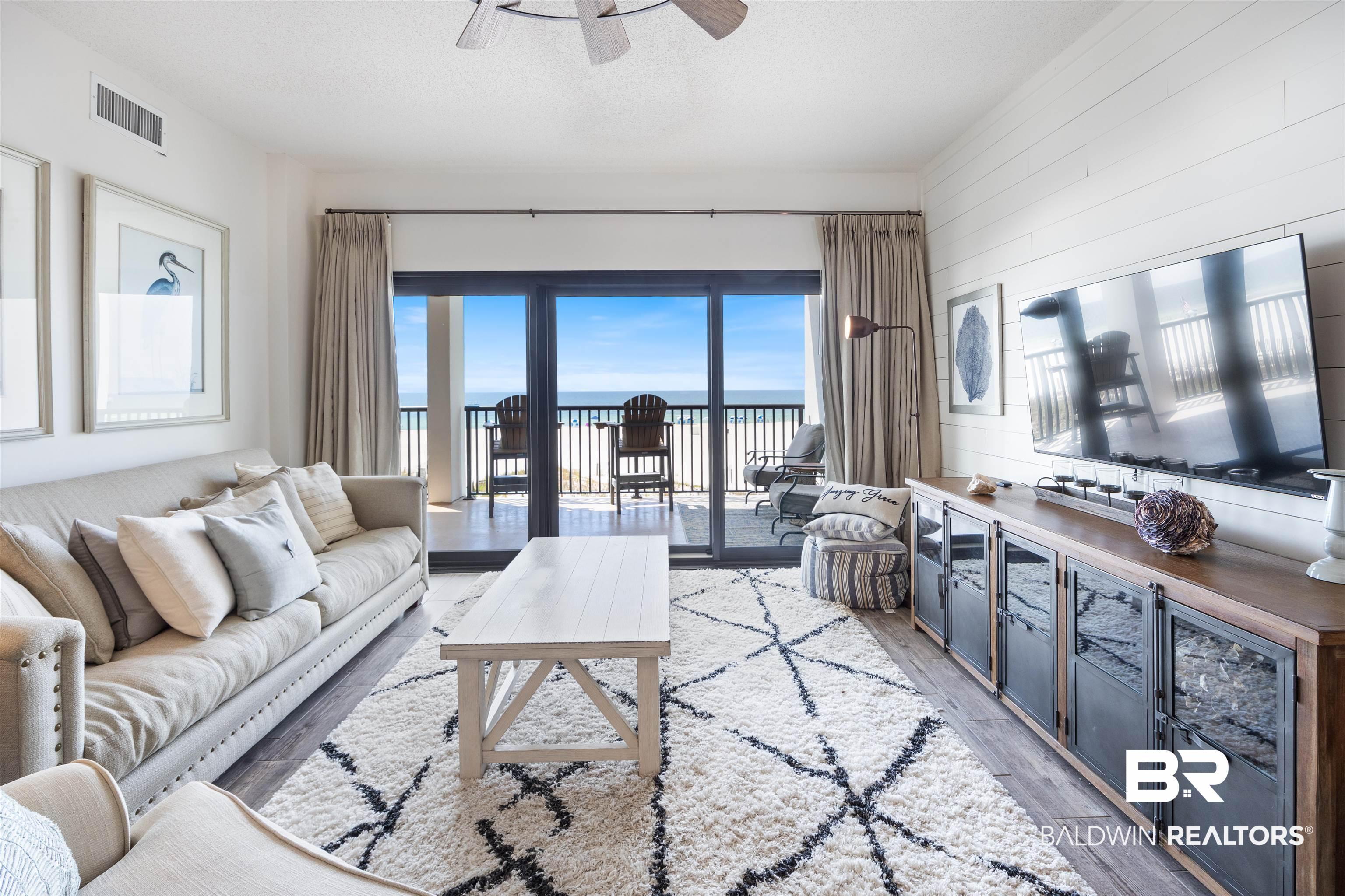 Image 2: Spacious living area featuring wood-finish flooring, a ceiling fan, and a wall of glass doors opening to a balcony with ocean scenery