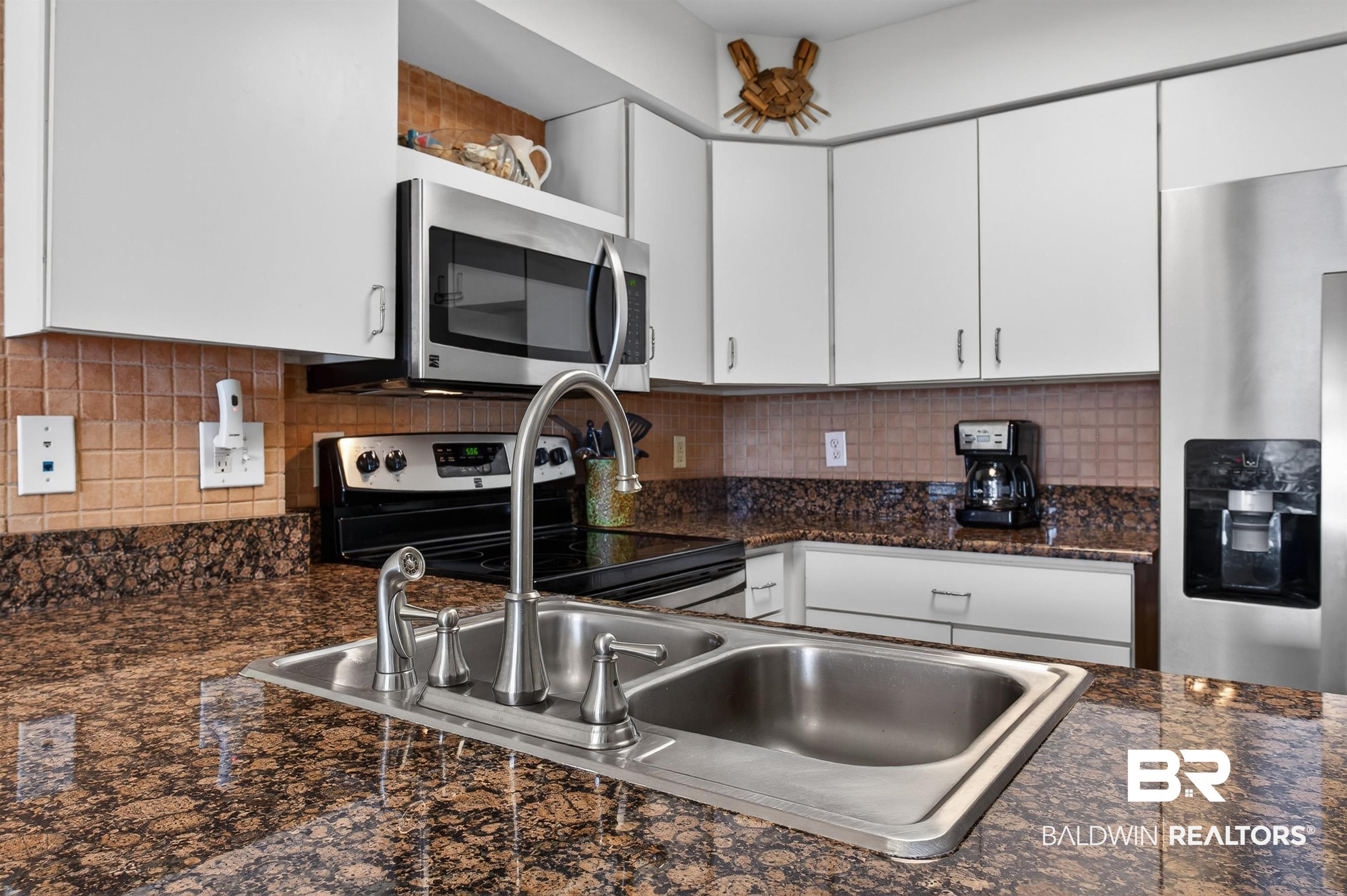 Image 4: Kitchen featuring a double-basin stainless steel sink with a high-arc faucet, granite countertops, tile backsplash, white cabinetry, and stainless steel appliances including a microwave, range, and refrigerator