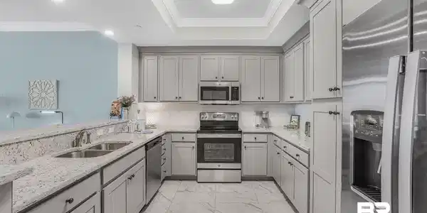 Image 2: U-shaped kitchen featuring light gray cabinetry with dark hardware, stainless steel appliances, a tile backsplash, and light-toned, speckled countertops