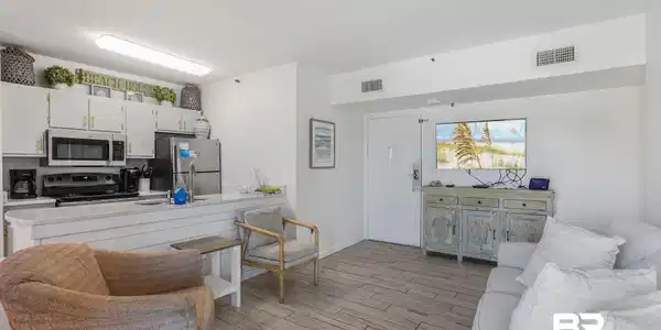 Image 3: Open-concept living area featuring wood-look tile flooring, white kitchen cabinetry with gold-tone hardware, stainless steel appliances including a microwave and refrigerator, a kitchen island with a sink, and recessed lighting