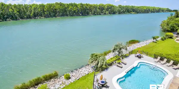 Image 3: Riverside property featuring a fenced swimming pool with a paved deck, mature palm trees, manicured lawns, and a riprap shoreline
