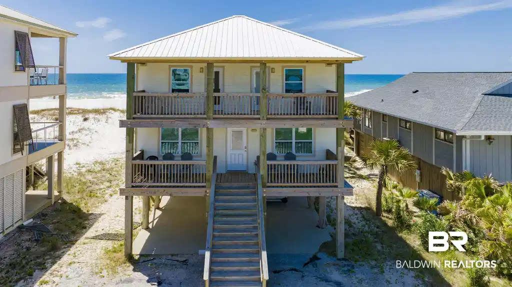 Image 1: Beachfront property featuring a white metal roof, two levels of covered balconies with wooden railings, and an elevated foundation over sandy terrain