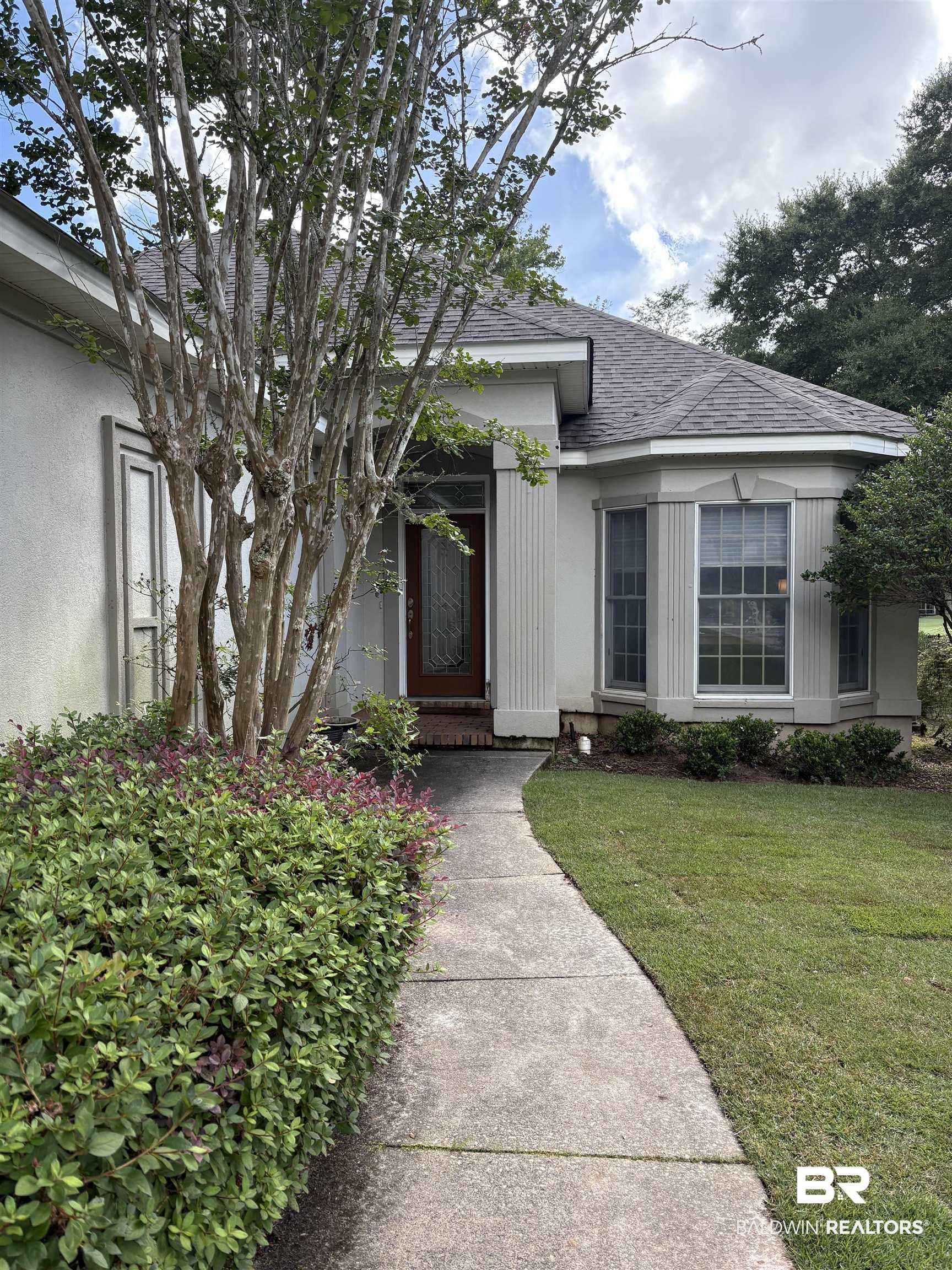 Image 1: Stucco exterior featuring a bay window, shingled roof, mature tree, concrete pathway, and lush landscaping