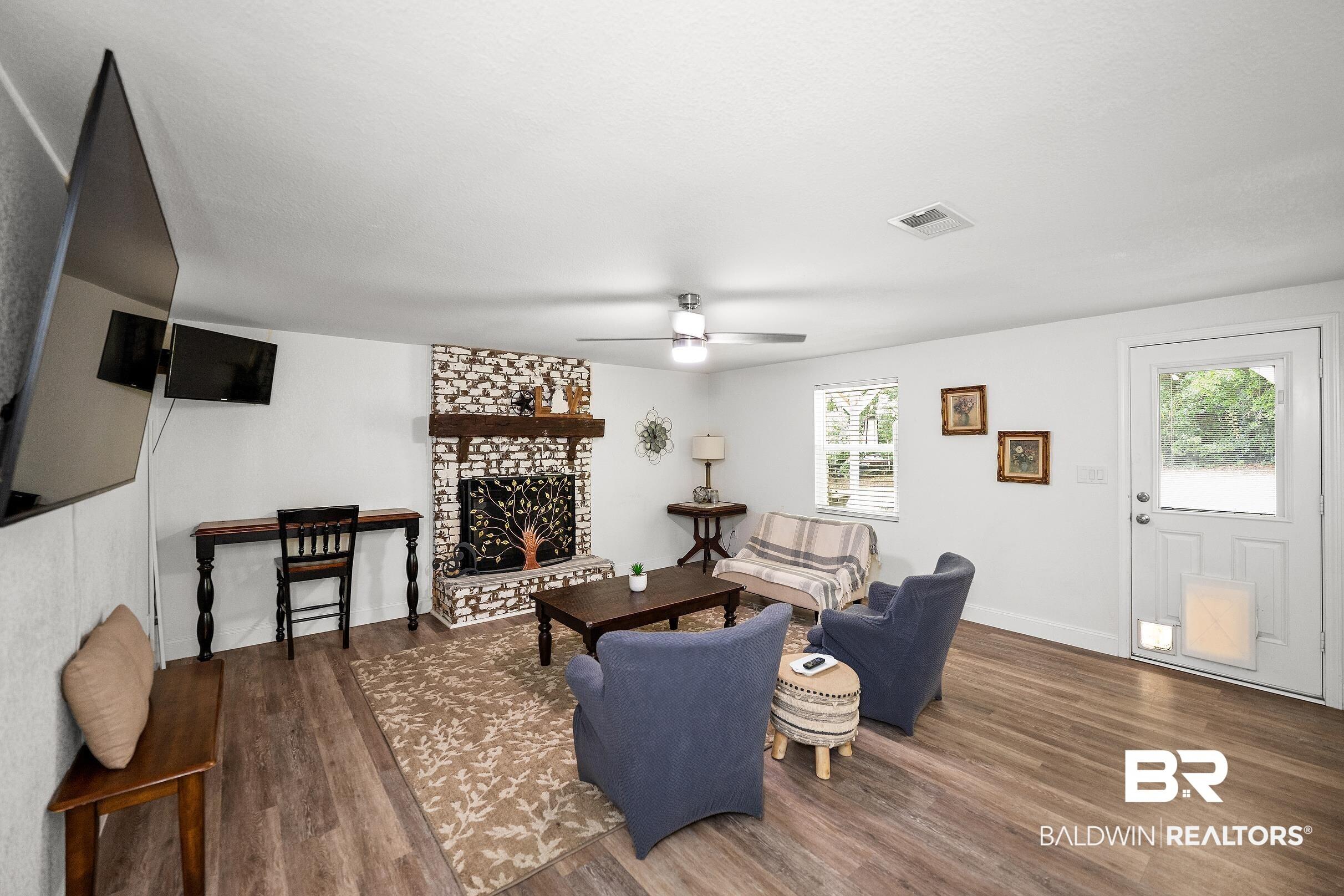 Image 4: Spacious living area featuring wood-finish flooring, a white brick fireplace with a wood mantel, and multiple windows providing natural light