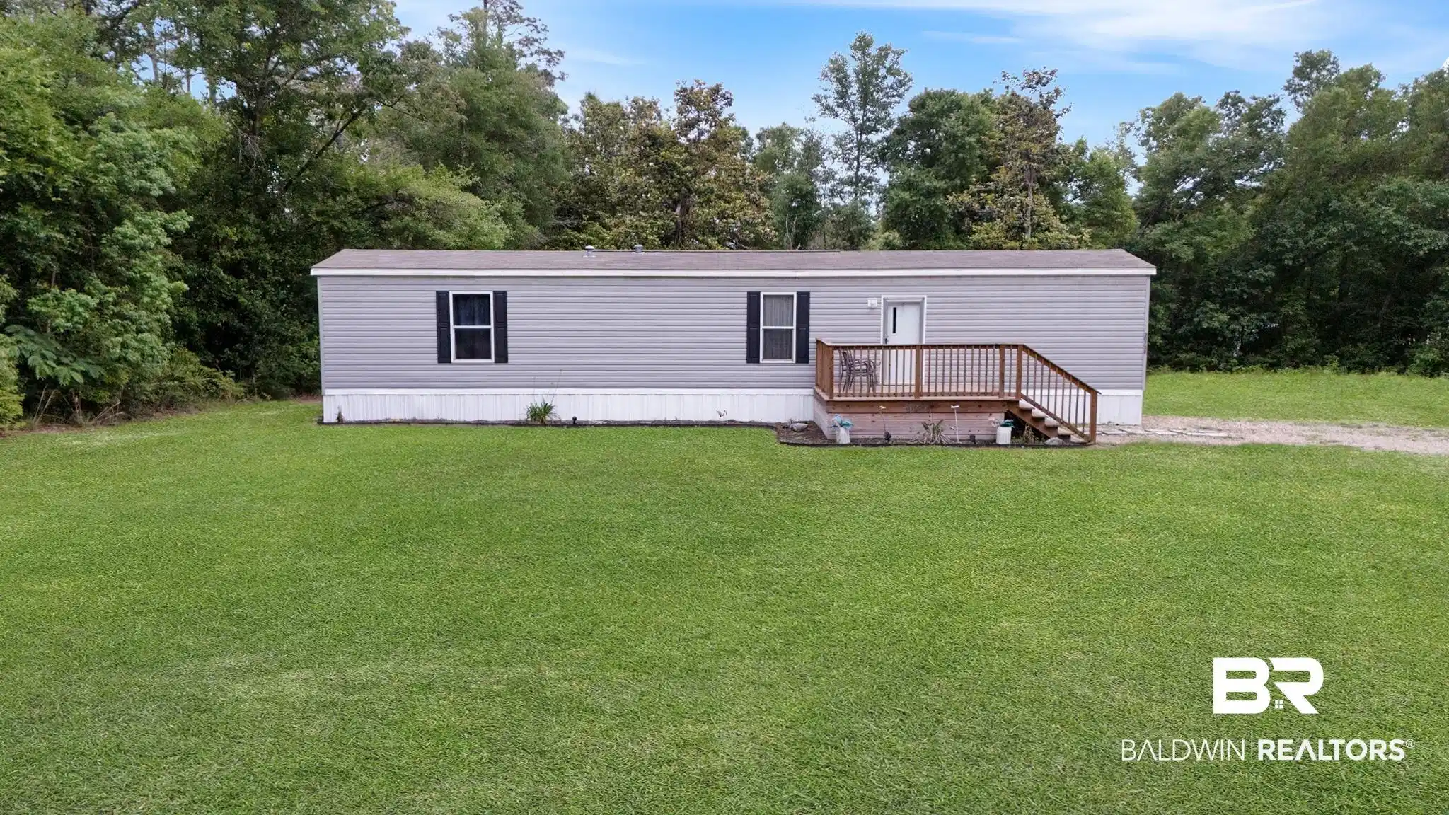 Image 1: Gray manufactured home with a brown wood deck, black shutters, and a white entry door