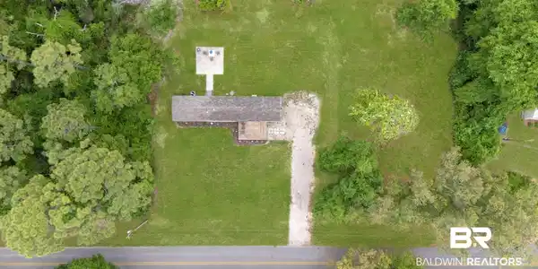 Image 4: Expansive green property featuring a single-story structure with a brown shingle roof, a gravel driveway, and a paved patio area