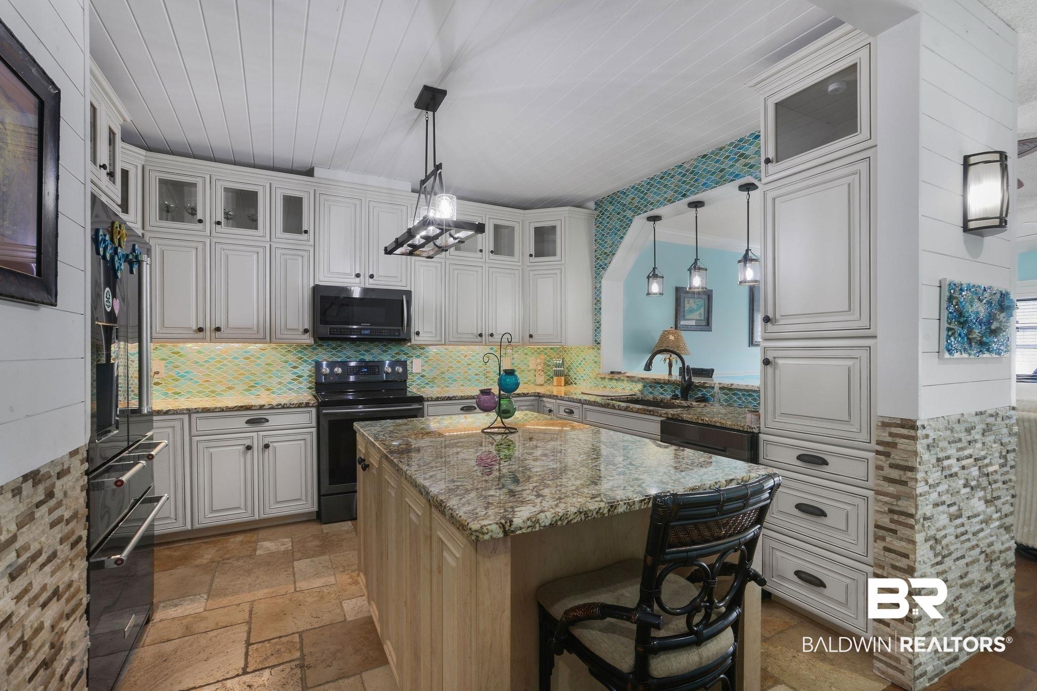 Image 3: Kitchen featuring white cabinetry, granite countertops, a tiled backsplash, a central island, and stone tile flooring