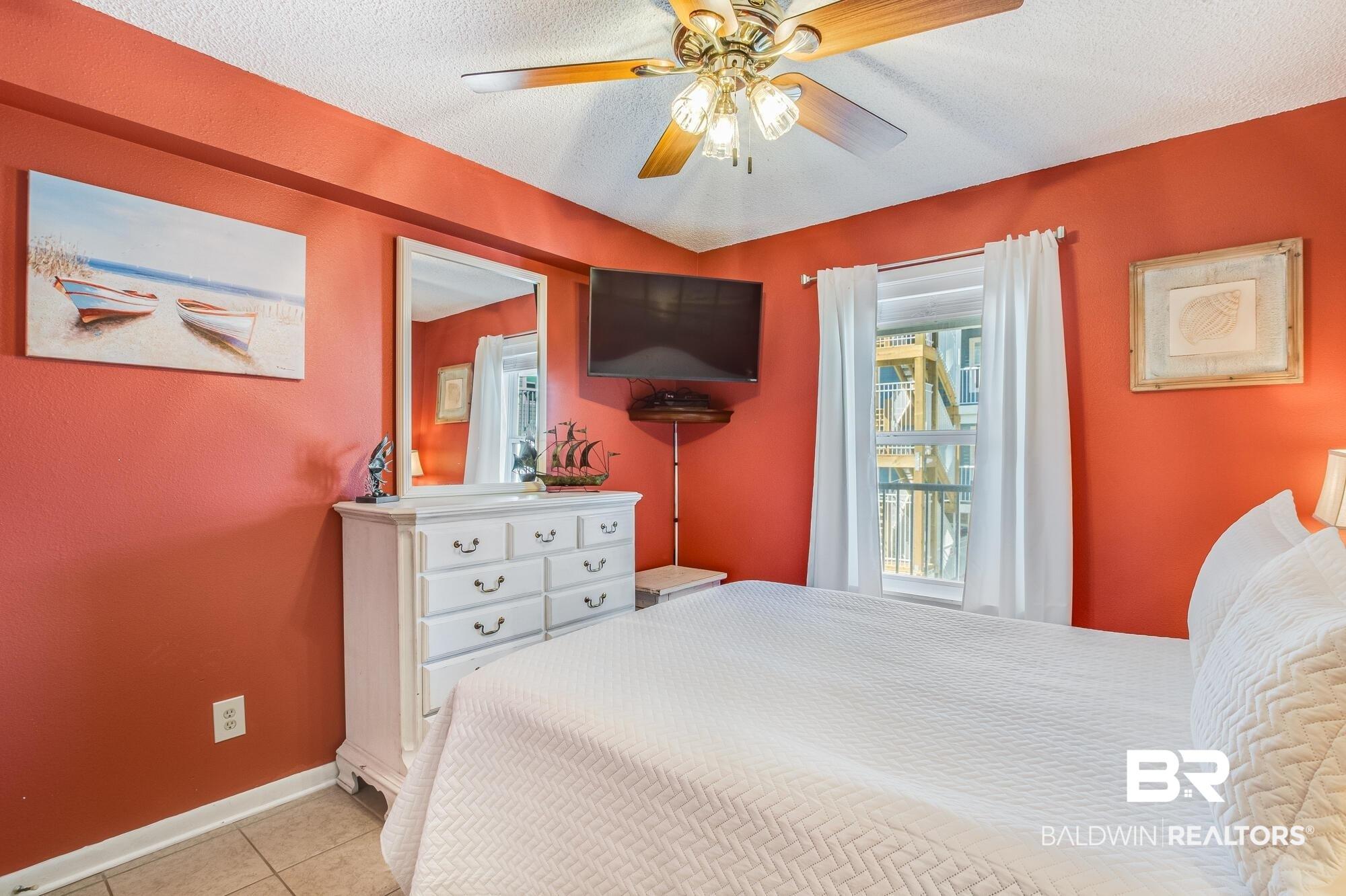 Image 4: Bedroom featuring a ceiling fan with integrated lighting, a wall-mounted television, and a window with white curtains