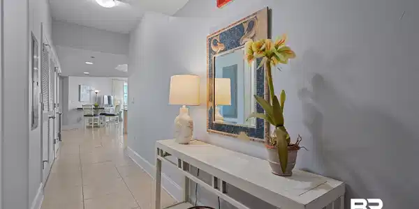 Image 3: Light-toned tile flooring extends through this hallway, featuring a decorative wall mirror, a console table, and louvered doors