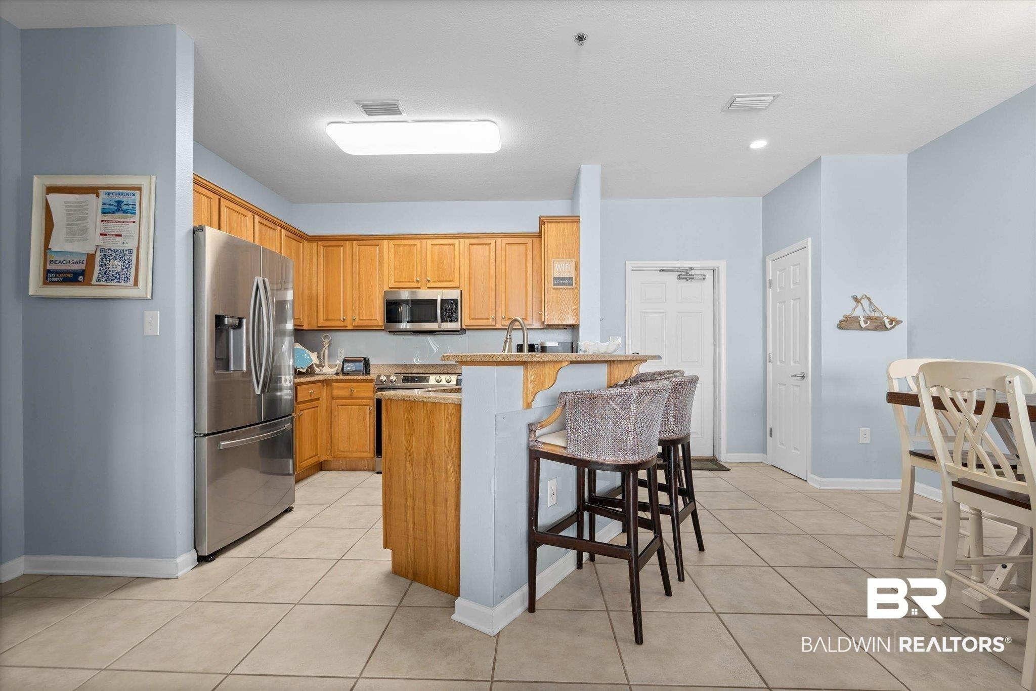 Image 2: Light-filled kitchen featuring stainless steel appliances, wood-finish cabinetry, neutral countertop surfaces, and tile flooring