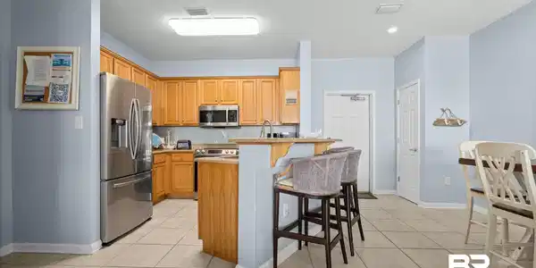 Image 2: Light-filled kitchen featuring stainless steel appliances, wood-finish cabinetry, neutral countertop surfaces, and tile flooring