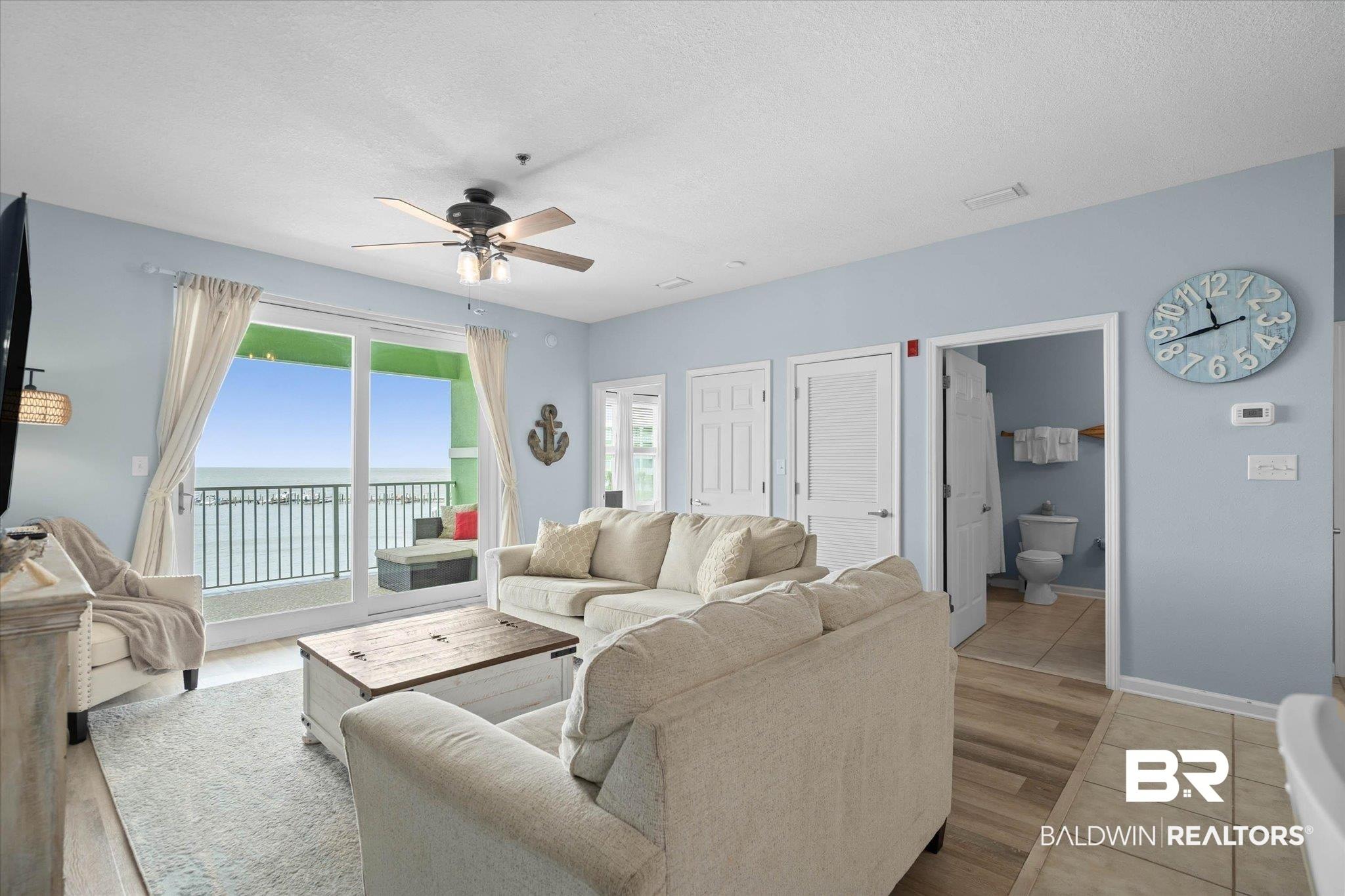 Image 4: Living area featuring expansive glass sliding doors opening to a balcony, wood-finish flooring, a ceiling fan with integrated lighting, and light blue wall paint