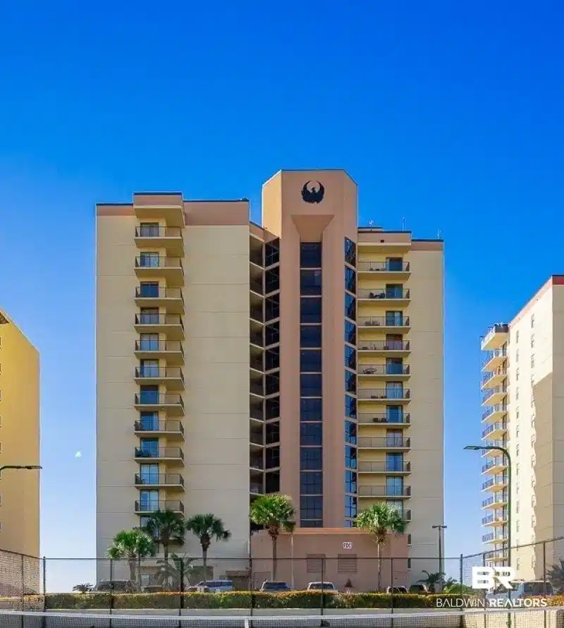 Image 1: Multi-story building featuring balconies with glass railings, a prominent central architectural element with a dark emblem, and a tennis court with a perimeter fence