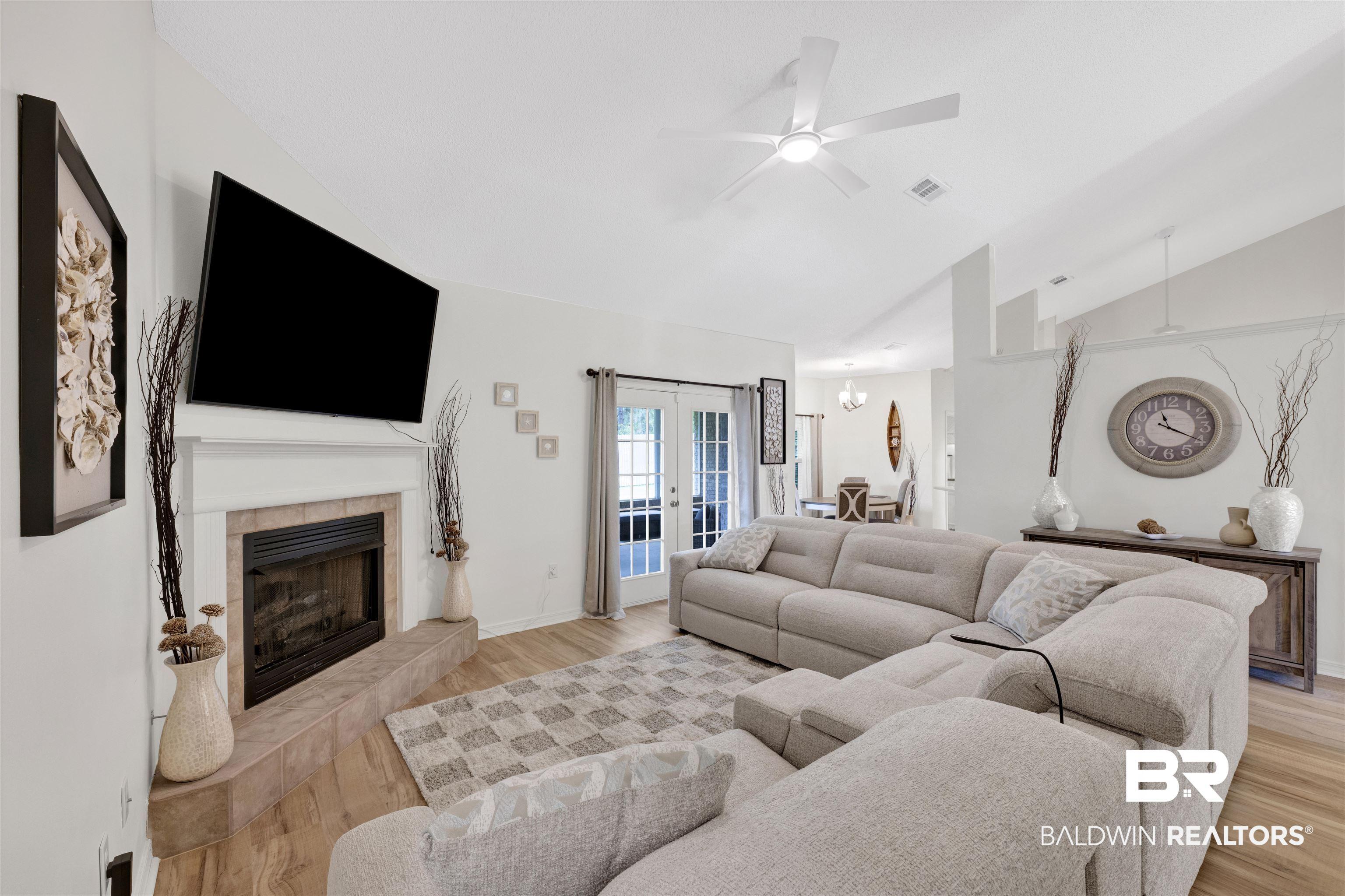 Image 4: Spacious living area featuring vaulted ceilings, wood-finish flooring, a fireplace with a tiled surround, and direct access to the outdoor space through double French doors