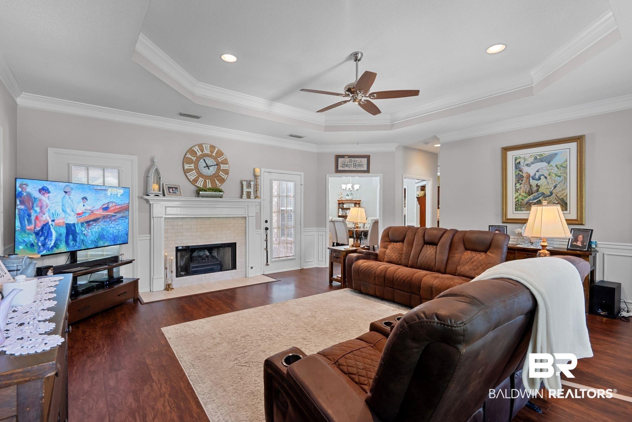 Image 4: Living area featuring wood-finish flooring, a tray ceiling with a ceiling fan, a fireplace with a tile surround and white mantel, crown molding, and wainscoting