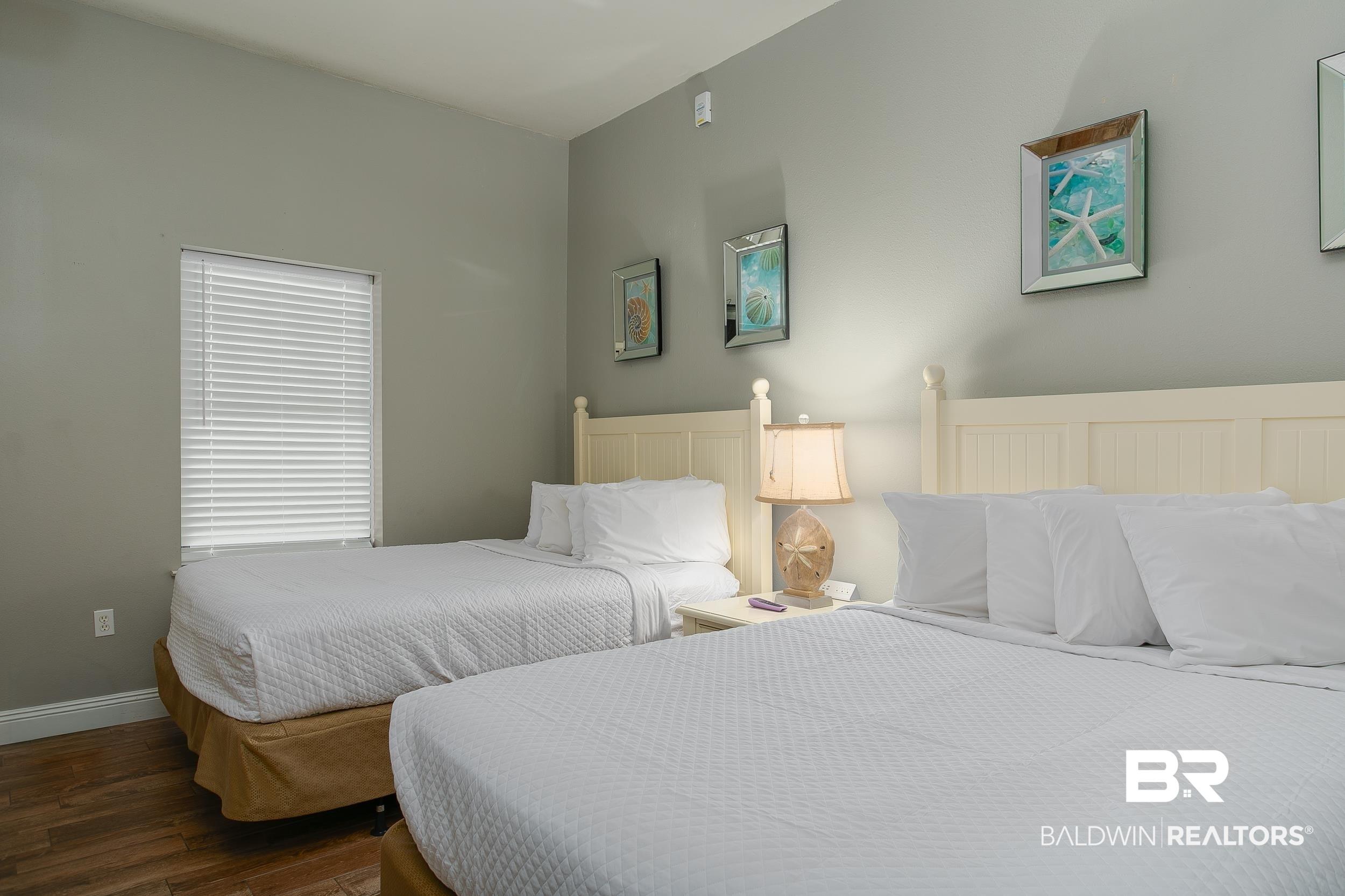 Image 2: Room featuring two beds with white headboards, a window with blinds, wood-finish flooring, and light grey walls