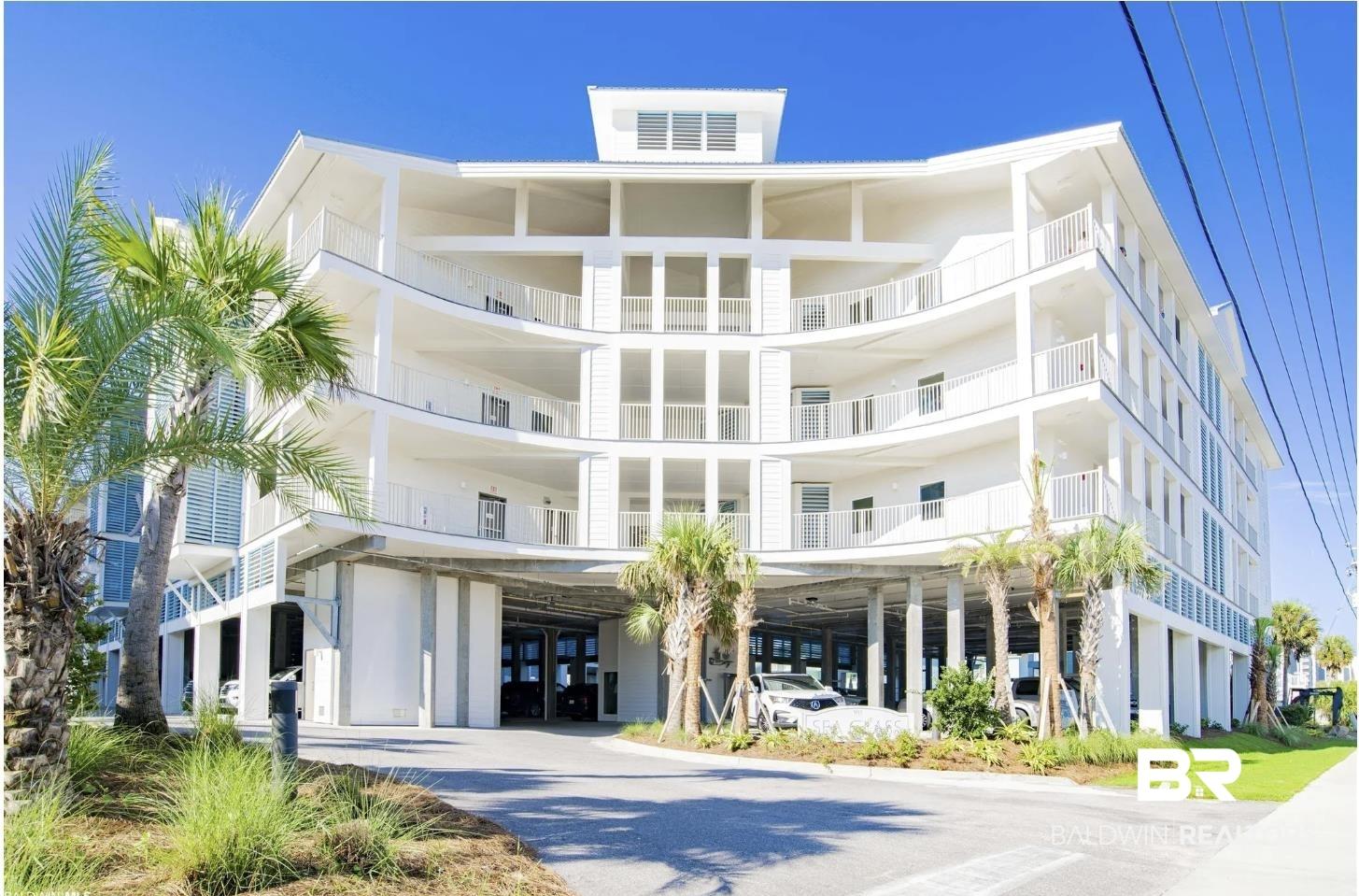 Image 1: Contemporary multi-story building featuring multiple balconies with white railings, a ground-level covered parking area, palm trees, and landscaped shrubbery