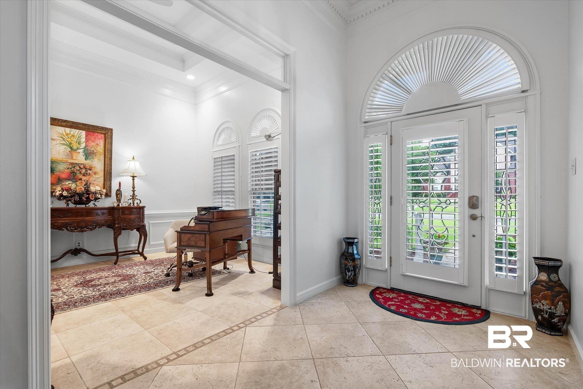 Image 4: Entryway featuring tile flooring with decorative inlay, a white paneled front door with decorative glass, two side panels with plantation shutters, and an arched transom window with plantation shutters