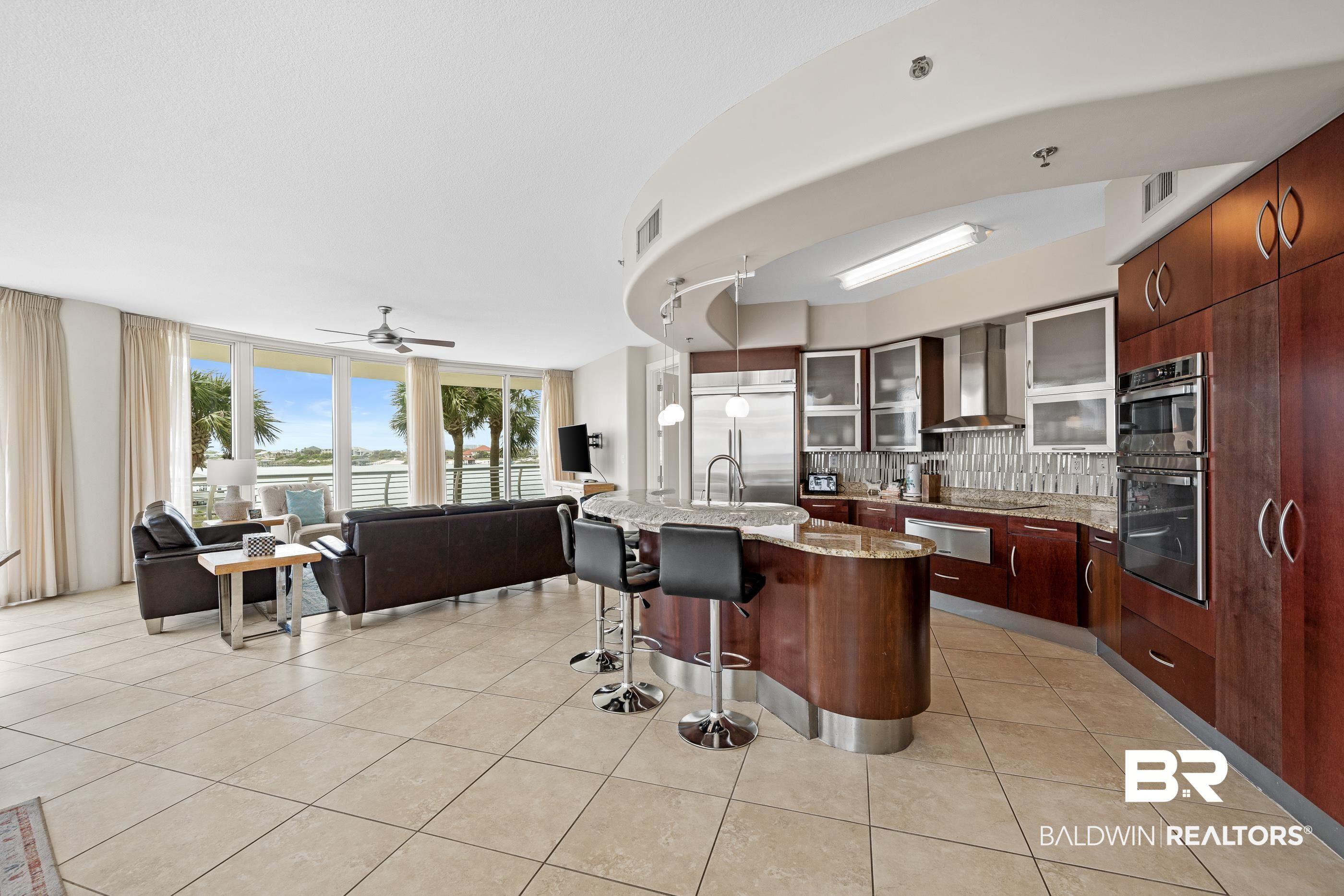 Image 3: Open-concept living space featuring expansive glass doors, light-toned tile flooring, and a modern kitchen with a curved island, integrated appliances, and wood-finish cabinetry