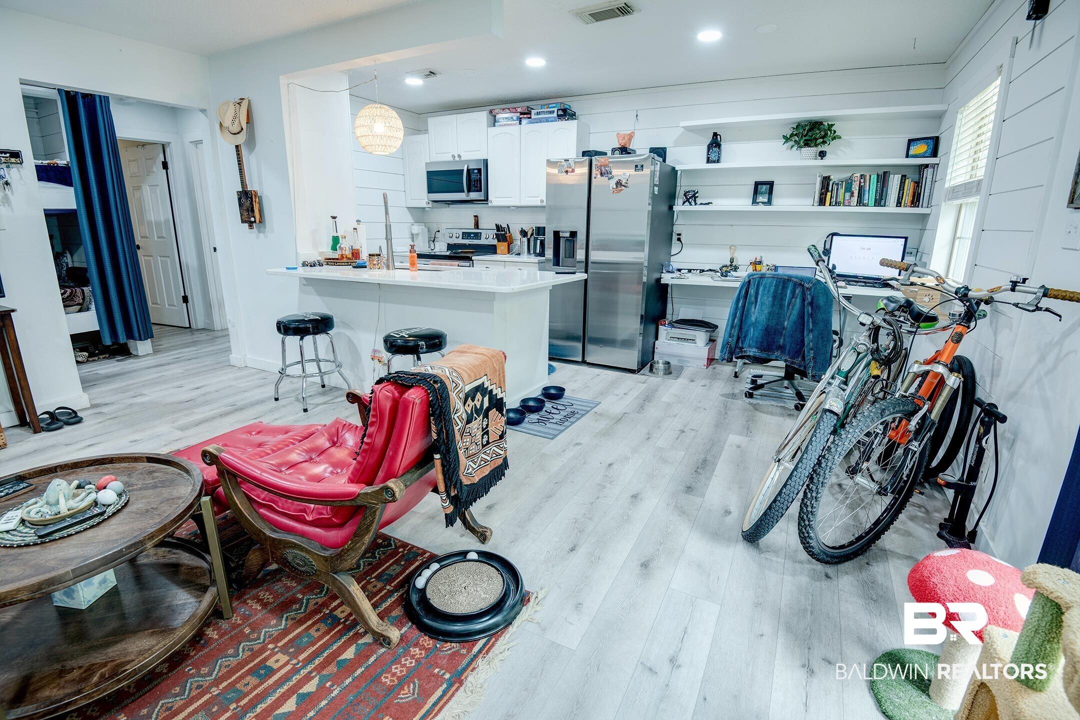 Image 4: Open-concept living area featuring light wood-finish flooring, white shiplap-style wall accents, a kitchen with white cabinetry and stainless steel appliances, a breakfast bar, and integrated shelving