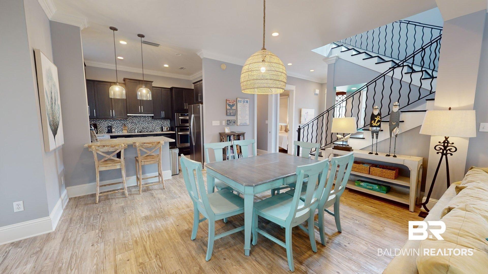 Image 4: Open-concept living space featuring wood-finish flooring, a contemporary kitchen with dark cabinetry and stainless steel appliances, a dining area with a woven pendant light, and a staircase with an ornamental wrought iron railing