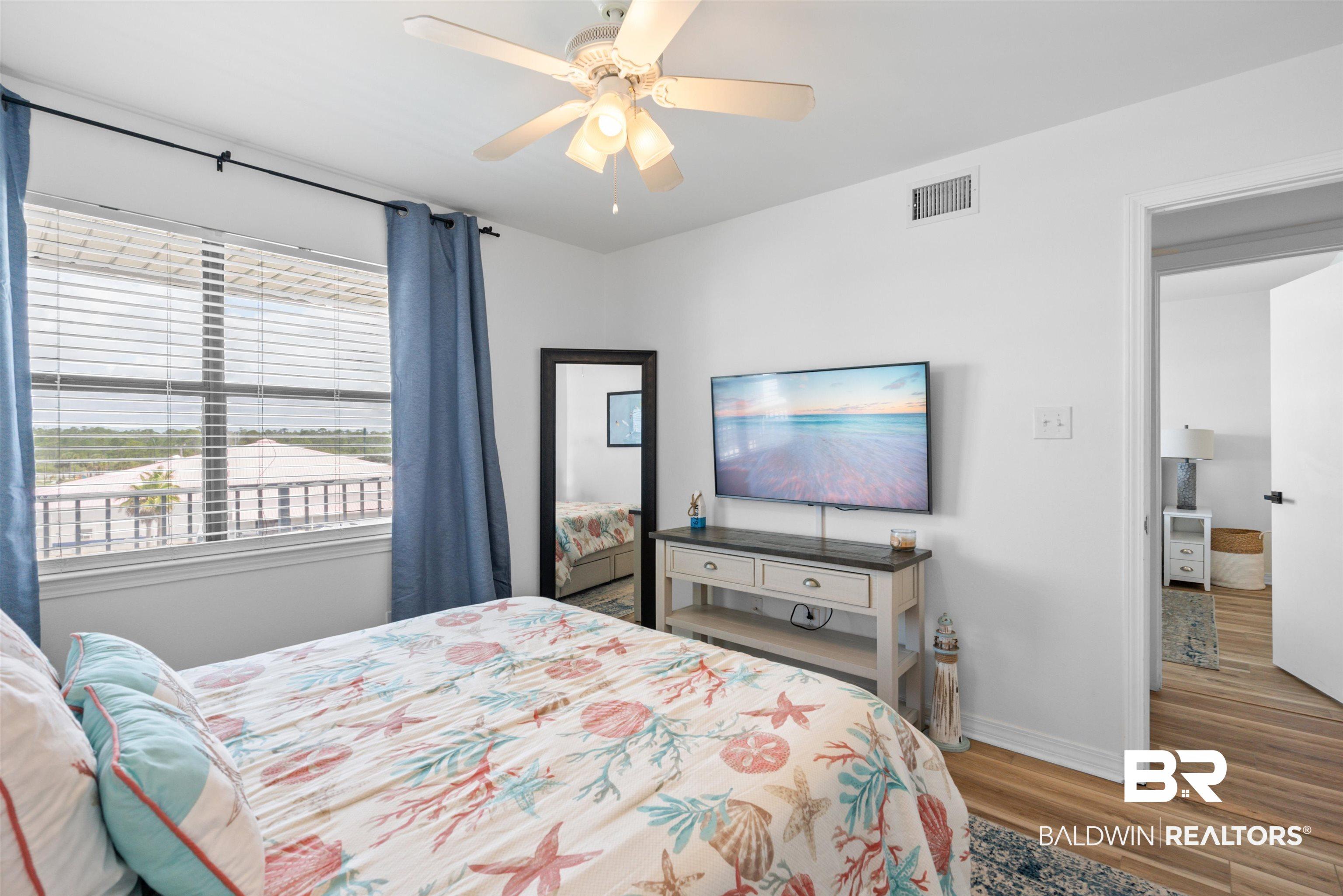Image 3: Bright bedroom featuring a large window with horizontal blinds, a ceiling fan with integrated lighting, wood-finish flooring, and white walls